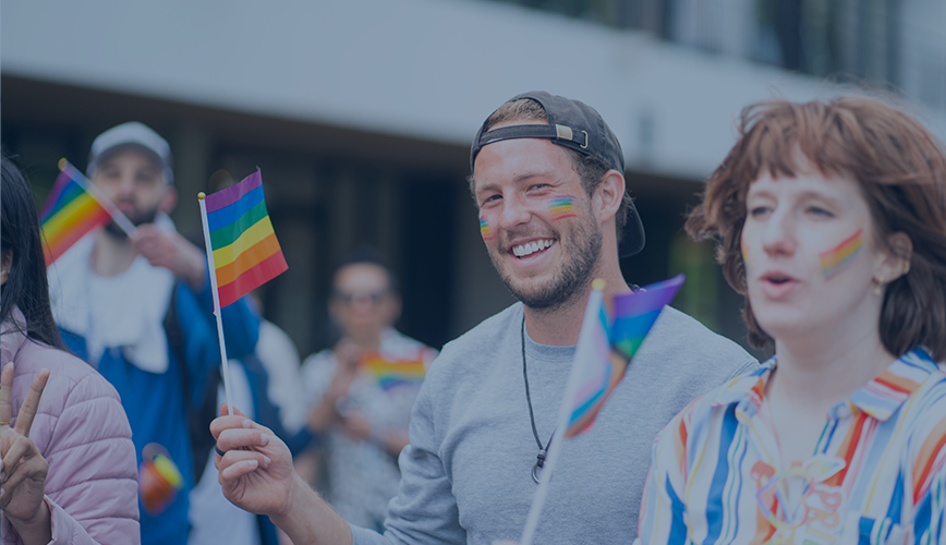 Deux personnes souriantes avec des drapeaux arc-en-ciel et des peintures faciales célébrant un événement LGBTQ+ sur le campus.