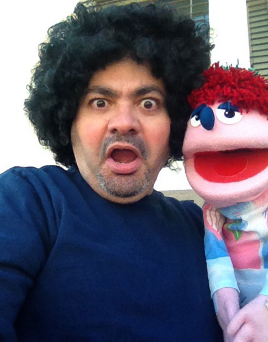 An expressive individual with a curly, dark-colored wig making a surprised facial expression while holding a colorful puppet or plush toy