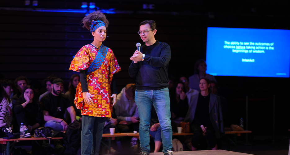 Two people stand on a stage during an event, with one person holding a microphone and speaking. The person speaking is dressed in casual clothing, while the other person is wearing a colorful traditional outfit. The audience is seated around the stage, attentively watching the presentation. A screen in the background displays a quote: 'The ability to use the outcomes of chosen futures before taking action is the beginning of wisdom. InterAct'