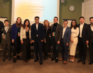 A group of twelve people posing together in front of a screen displaying 'HEC Paris Le Château'. They are all dressed in business or smart casual attire, smiling and standing in a row. The group includes both men and women of diverse backgrounds