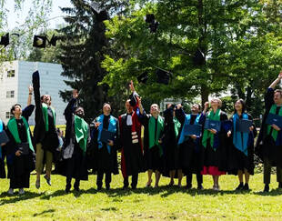 A group of doctoral candidates in graduation gowns and hoods celebrating outdoors by throwing their caps into the air. They are standing on a grassy area surrounded by trees and a building in the background. The graduates are smiling and holding their diplomas as they commemorate this significant milestone.